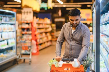 Parlak bir markette taze meyve ve sütle dolu turuncu bir alışveriş arabasını iten adam.