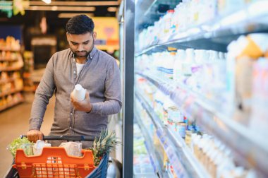 Markette alışveriş yapan genç bir adam, dondurulmuş süt reyonundaki süt şişesindeki etiketleri karşılaştırıyor.