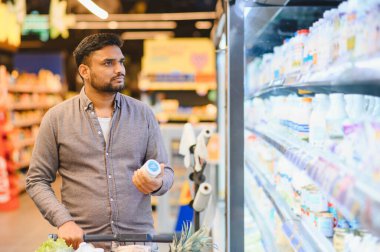 Elinde yoğurt tutan ve alışveriş arabasını iten bir adam marketin dondurulmuş süt ürünlerini arıyor.