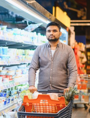 Süpermarkette alışveriş yaparken alışveriş arabasını iten bir adam, alışveriş arabasını taze yiyeceklerle dolduruyor.