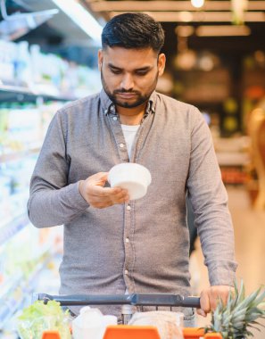 Bir ürünü tutan ve etiketini dikkatlice okuyan bir adam parlak ışıklı bir markette bir alışveriş arabasını iterken...