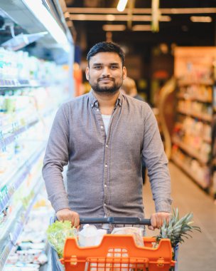 Genç adam market alışverişi yapıyor, taze meyve ve sebzelerle dolu bir alışveriş arabasını parlak ışıklı bir süpermarkette itiyor.