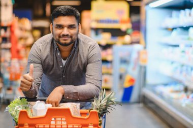 Süpermarket koridorunda içi taze yiyeceklerle dolu bir alışveriş arabasını iten bir adam satın almasından memnun olduğunu gösterir.