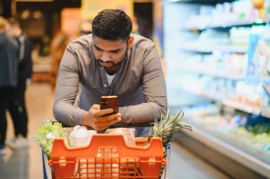 Marketten yiyecek alırken akıllı telefonunu dijital alışveriş listesini kontrol etmek için kullanan bir adam.