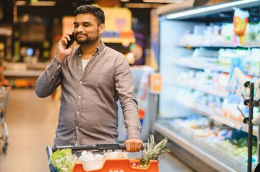 Gülümseyen bir adam alışveriş arabasını itiyor, market koridorunda telefonla konuşuyor.