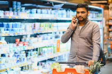 Market koridorlarında gezinen genç adam iletişim için akıllı telefon kullanıyor ya da alışveriş listesini kontrol ediyor, alışveriş arabası itiyor.