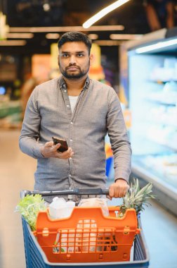 Perakende mağazasından alışveriş yapan bir adam, içi taze yiyecek dolu bir arabayı iterken akıllı telefonu kontrol ediyor.