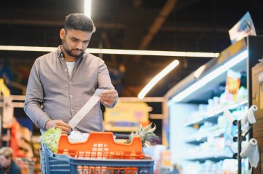 Elinde uzun bir alışveriş makbuzu olan adam, alışveriş arabasıyla bakkal koridorunda dikilirken fiyatları inceliyor.