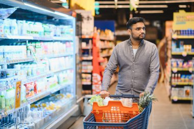 Taze ürünlerle dolu bir alışveriş arabasını iten bir adam ve parlak bir süpermarket koridorunda paketlenmiş ürünler.