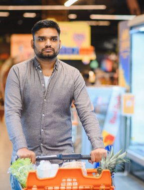 Süpermarket koridorunda içi taze ürünlerle dolu turuncu alışveriş arabalarını iten adam sağlıklı sebzeleri seçmeye odaklanmış.