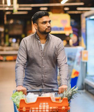 Genç adam market alışverişi yapıyor, içi taze ürünlerle dolu bir arabayı süpermarket koridorunda itiyor.