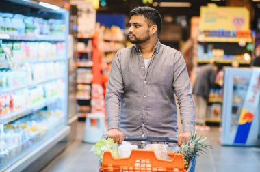 Genç adam alışveriş yapıyor, markette soğuk bir koridorda turuncu bir arabayı itiyor, bakkaliye arıyor.