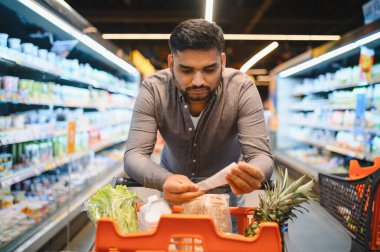 Market koridorunda alışveriş listesinde eşyalar bulan adam, içi taze ürün ve ekmekle dolu bir tramvayı itiyor.