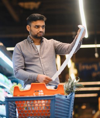 Süpermarketteki genç adam, uzun ve pahalı faturaları alışveriş arabasındaki eşyalarla karşılaştırıyor, gıda ve bütçe artışından endişe ediyor.