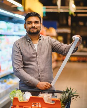 Süpermarkette elinde uzun bir makbuz tutan bir adam alışveriş yaparken artan yaşam ve tüketim maliyetlerini gösteriyor.