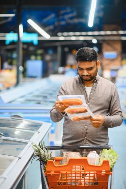 Dondurulmuş somon fileto paketlerini elinde tutan bir adam market koridorunda elinde alışveriş arabasıyla ürün bilgilerine bakıyor.