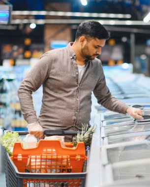 Eldiven giyen bir müşteri buzdolabı vitrininden eşyalar seçiyor, perakende mağazasında içi yiyecek dolu bir alışveriş arabasını itiyor.