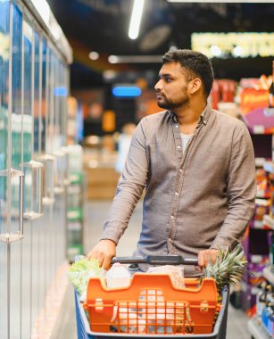 Market alışverişi yapan genç bir adam. Parlak bir süpermarket koridorunda dolu bir alışveriş arabasını iten bir adam.