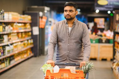 Genç adam market alışverişi yapıyor, içinde yiyecek olan bir arabayı modern bir süpermarkette koridorda itiyor.