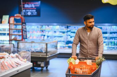 Genç adam market alışverişi yapıyor, taze ürünlerle dolu bir arabayı modern bir markette itiyor.