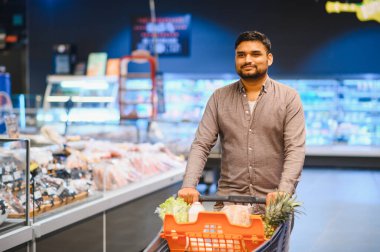 Market alışverişi yapan genç bir adam, modern bir süpermarketin taze gıda reyonunda dolu bir arabayı iterek...