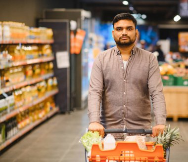 Market alışverişi yapan, alışveriş arabasını koridorda iten, kameraya bakan genç bir adam. Tüketim ve perakende kavramı