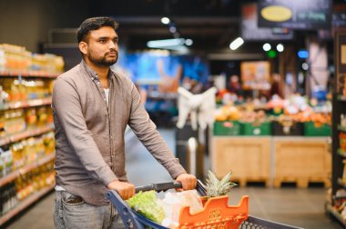 Genç bir adam marketin koridorunda içi taze ürünlerle dolu bir market arabasını itiyor. Seçeneklere odaklanıyor.