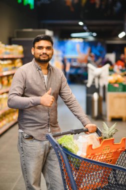 Alışverişle dolu bir alışveriş arabasını iten adam, normal bir süpermarket gezisi sırasında memnuniyet ve onay gösteriyor.