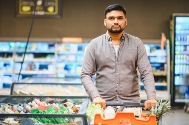Market alışverişi yapan genç bir adam. Parlak ışıklı bir süpermarkette içi taze ürünler ve gerekli malzemelerle dolu bir arabayı itiyor.