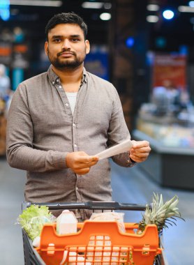 Genç adam alışveriş listesindeki eşyaları kontrol ederken markette içi taze ürünlerle dolu bir arabayı itiyor.