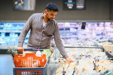 Market alışverişi yapan genç bir adam, modern bir süpermarketin vitrininden taze peynir seçiyor.