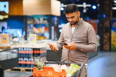 Süpermarkette elinde alışveriş arabasıyla genç sakallı bir adam, alışveriş yaparken ve seçeneklerini karşılaştırırken akıllı telefon uygulamalarını kontrol ediyor.
