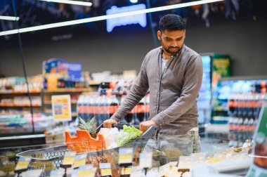 Market koridorunda ürünleri kontrol ederken market arabasını iten adam. Tüketim ve günlük alışveriş kavramı