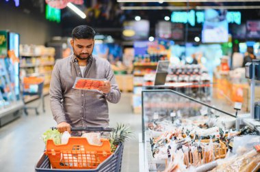 Genç adam modern bir süpermarkette alışveriş yapıyor, elinde bir paket taze et tutuyor, market alışverişi için tüketici seçimi yapıyor.
