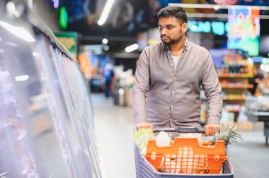 Yüklü bir alışveriş arabasını iten, süpermarkette dondurulmuş gıda reyonuna göz gezdiren, yiyecek seçen tüketici.