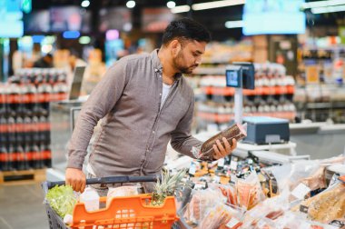 Marketin et ve meze bölümünde paketlenmiş salam ya da sosisi incelerken alışveriş arabasını iten adam.