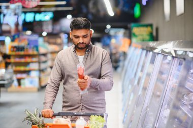 Müşteri soğutuculu vitrinden paketlenmiş et ürünü seçiyor, içi yiyecek dolu bir alışveriş arabasını itiyor.