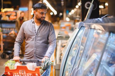 Hintli bir adam alışveriş arabasını itiyor, marketin soğutmalı bölümünde sergilenen yiyeceklere bakıyor.