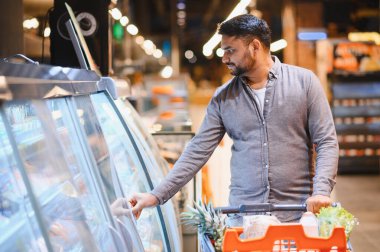 Süpermarket koridorunda alışveriş yapan bir adam, bir alışveriş arabasını iterken soğutuculu vitrin açıyor.