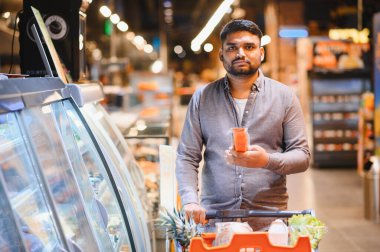 Genç adam süpermarketten alışveriş yapıyor, elinde bir ürün tutuyor ve alışveriş arabasıyla alışveriş yapıyor. Tüketim ve sağlıklı yaşam tarzı kavramı