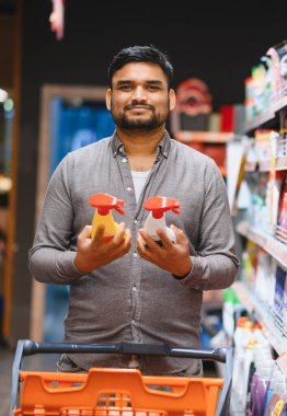 Süpermarket koridorunda elinde iki temizlik spreyi tutan adam, ev hijyeni ürünleri hakkında karar veriyor.