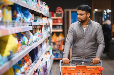 Genç bir adam süpermarket koridorunda turuncu bir alışveriş arabasını itiyor, raflardan ürünleri seçiyor.
