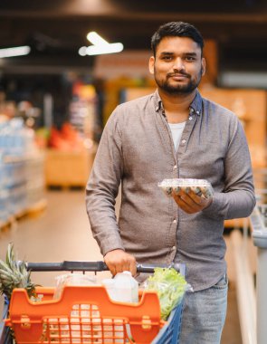 Bakkal dolu bir alışveriş arabasını iten, bıldırcın yumurtası taşıyan, modern bir süpermarkette gezinen bir adam.