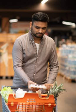 Modern bir süpermarket koridorunda içi yiyecek ve yiyecek dolu bir alışveriş arabasını iten bir adam.