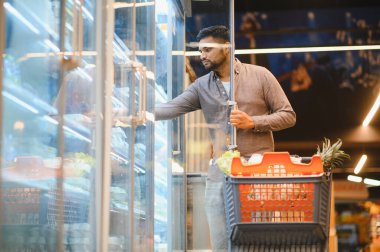 Bir markette dondurucu bölümünden yiyecek seçen bir adam, dolu bir alışveriş arabasını itiyor. Günlük alışveriş rutini