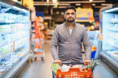 Parlak bir süpermarket koridorunda içi taze ürünlerle dolu bir alışveriş arabasını iten adam doğrudan kameraya bakıyor.