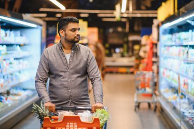 Market alışverişi yapan genç bir adam. Parlak ışıklı bir süpermarket koridorunda içi yiyecek dolu turuncu alışveriş arabalarını itiyor.