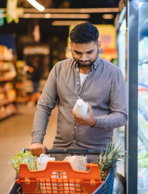 Elinde bir ürün şişesi olan genç bir adam, markette alışveriş yaparken seçenekleri karşılaştırıyor.