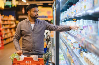 Bir marketteki soğutuculu raftan süt ürünü seçen bir adam içi yiyecek dolu bir alışveriş arabasını itiyor.