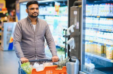 Genç Hintli bir adam markette içi yiyecek dolu bir alışveriş arabasını itiyor ve yeni ürünler arıyor.